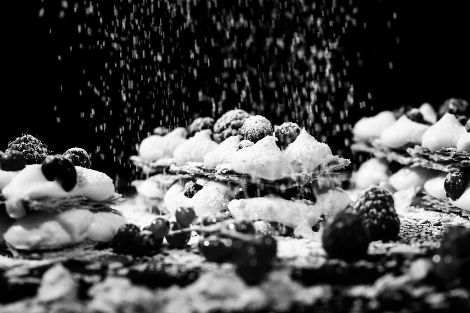 Black and white photo of pastries dusted with powdered sugar and topped with blackberries on a dark background.