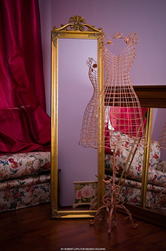Ornate gold floor mirror beside a wire dress form with pink lamp in a room with floral furniture.