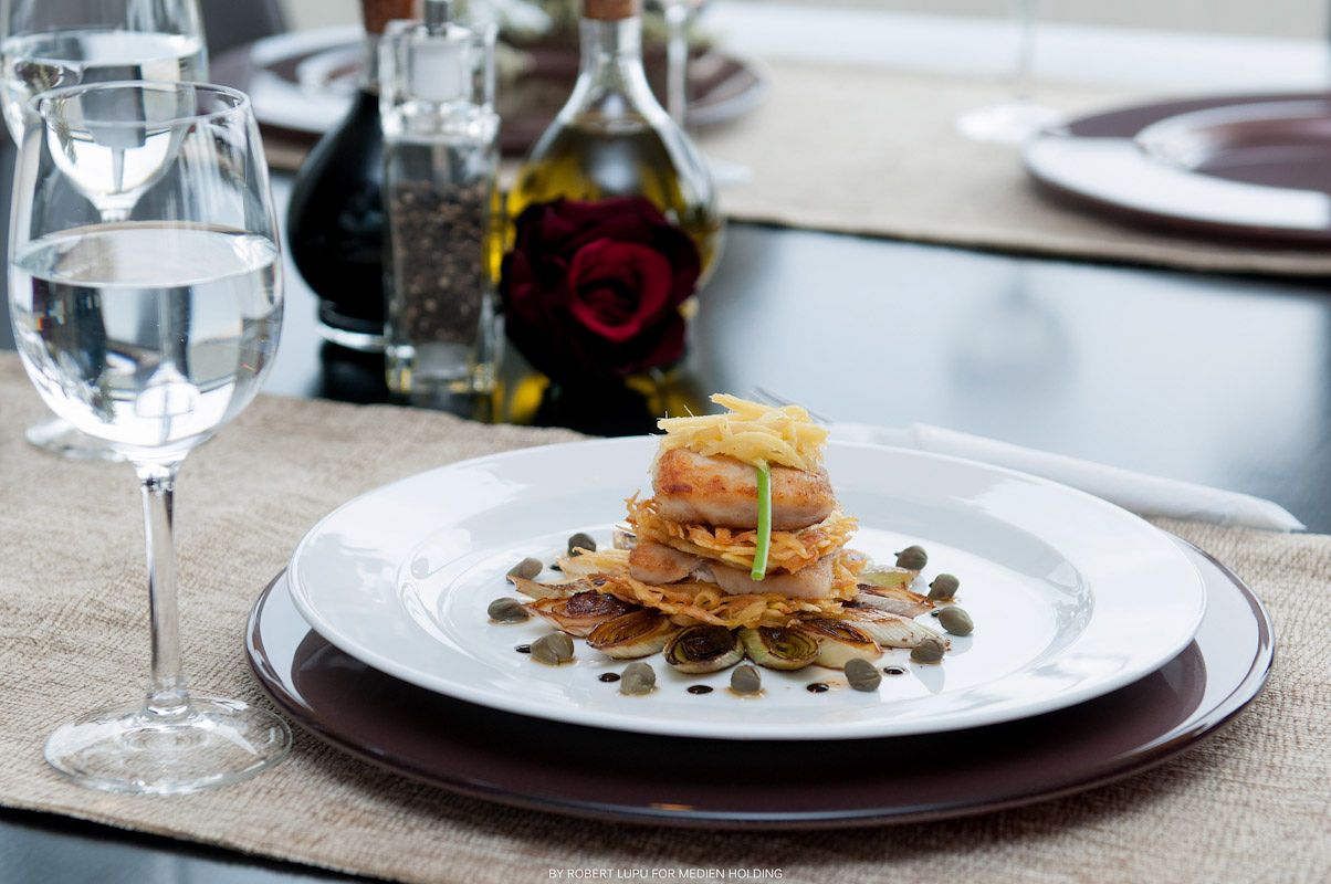 Elegant gourmet dish stacked on white plate with seeds, garnished herbs, wine glass and oil bottle in background.
