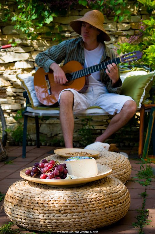Man in straw hat playing acoustic guitar outdoors, with grapes and cheese on woven ottoman in garden setting.