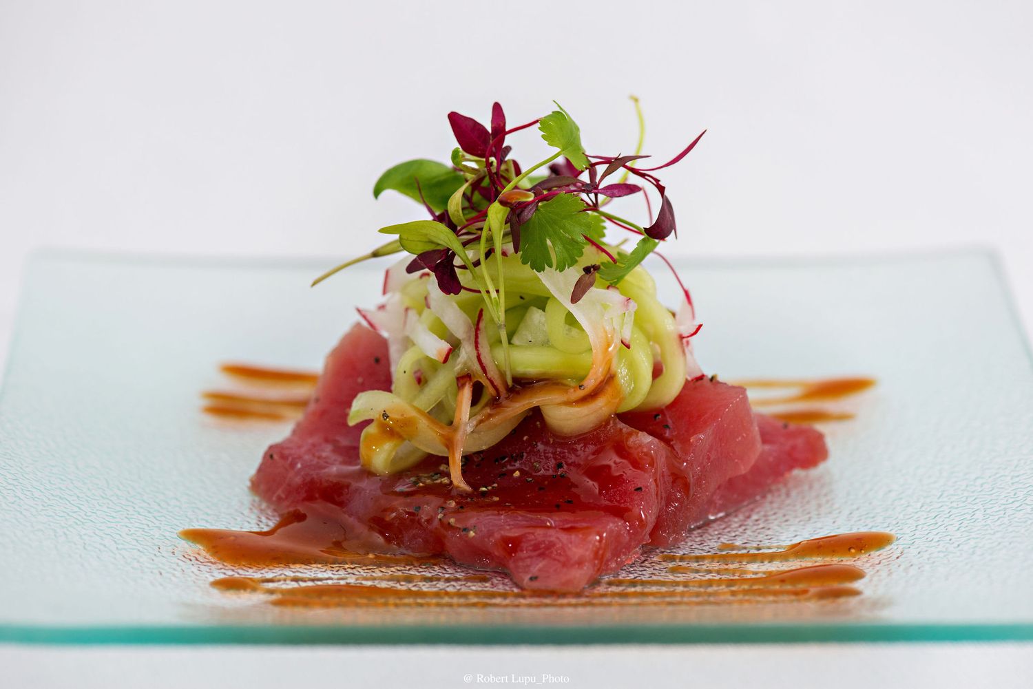 Fresh tuna tartare topped with microgreens and avocado, elegantly plated on a square glass dish with sauce drizzle.