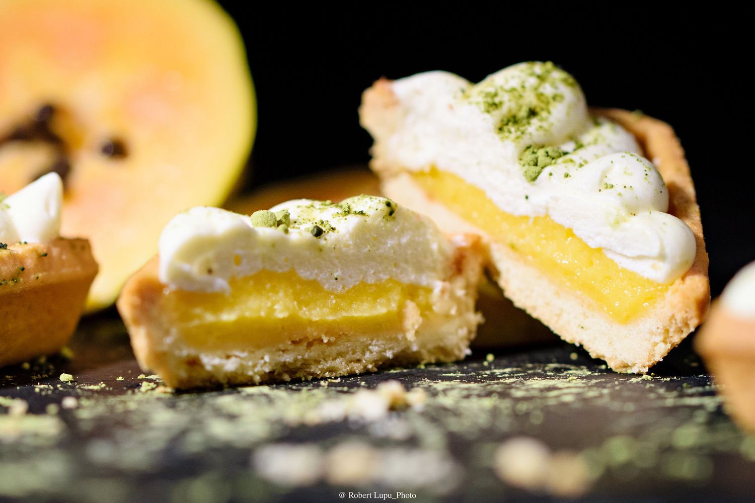 Close-up of sliced lemon tart with creamy meringue topping and yellow curd filling on dark surface.