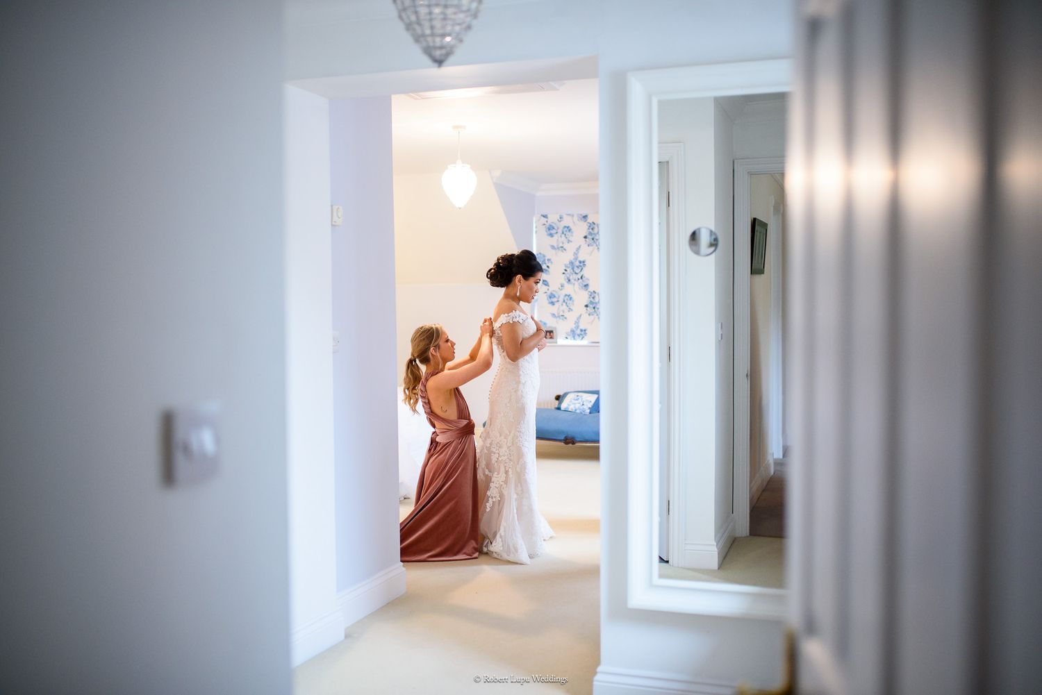 Bridesmaid in pink dress helping bride with wedding gown in a bright doorway, with mirror reflection visible.