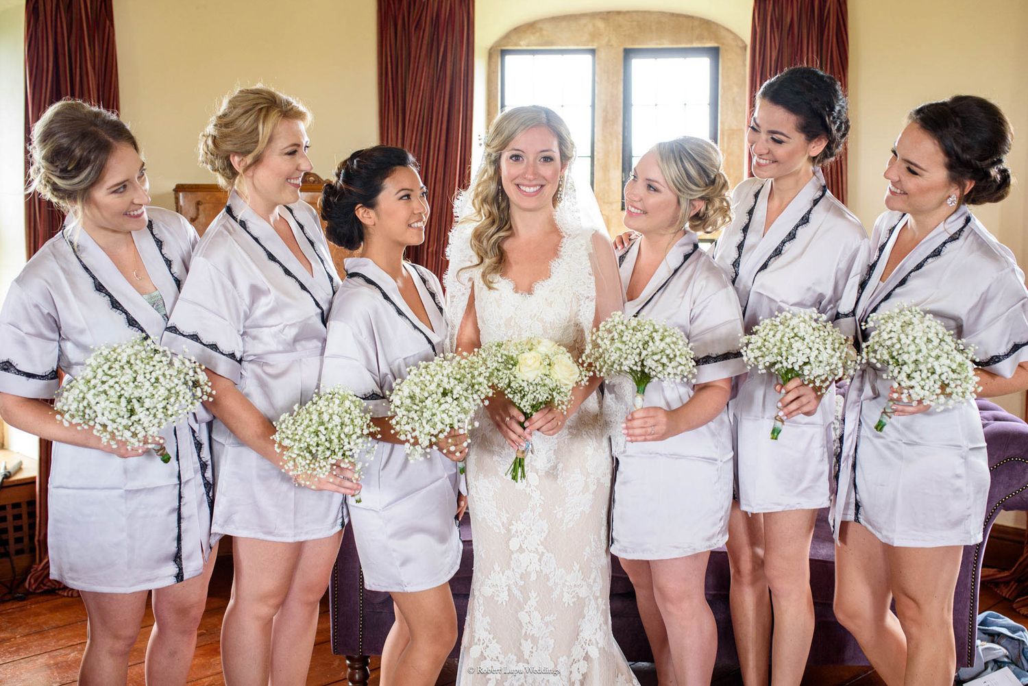Bride in lace gown poses with six bridesmaids in matching gray robes holding white baby's breath bouquets indoors.