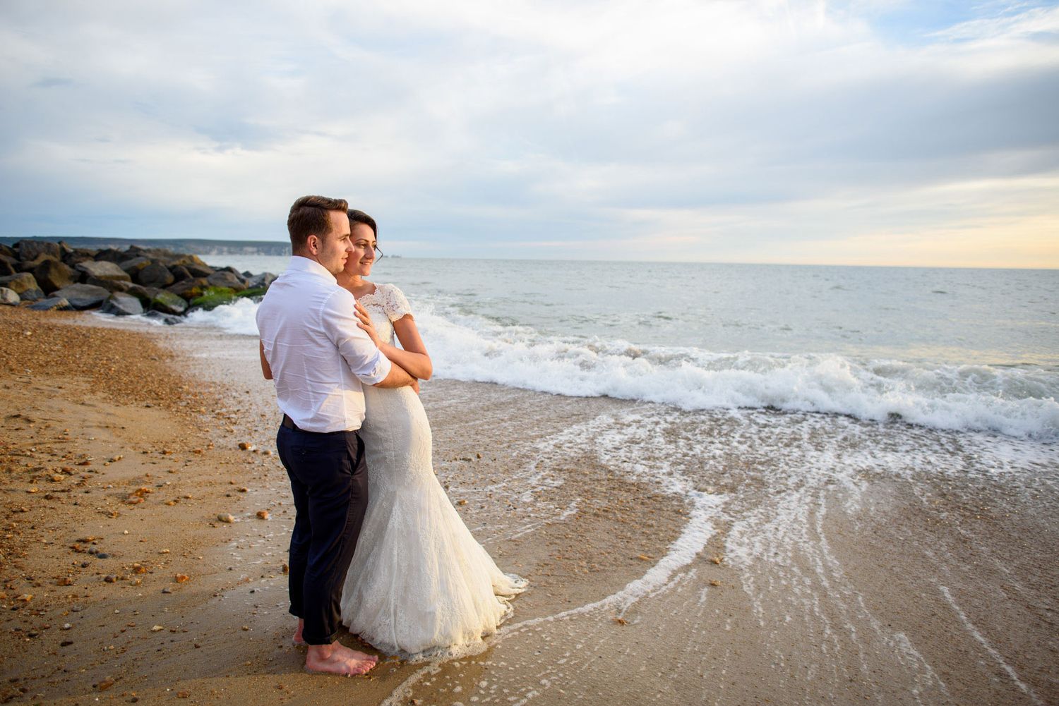 A couple in wedding attire embraces on a sandy beach at sunset with ocean waves and rocky jetty behind them.