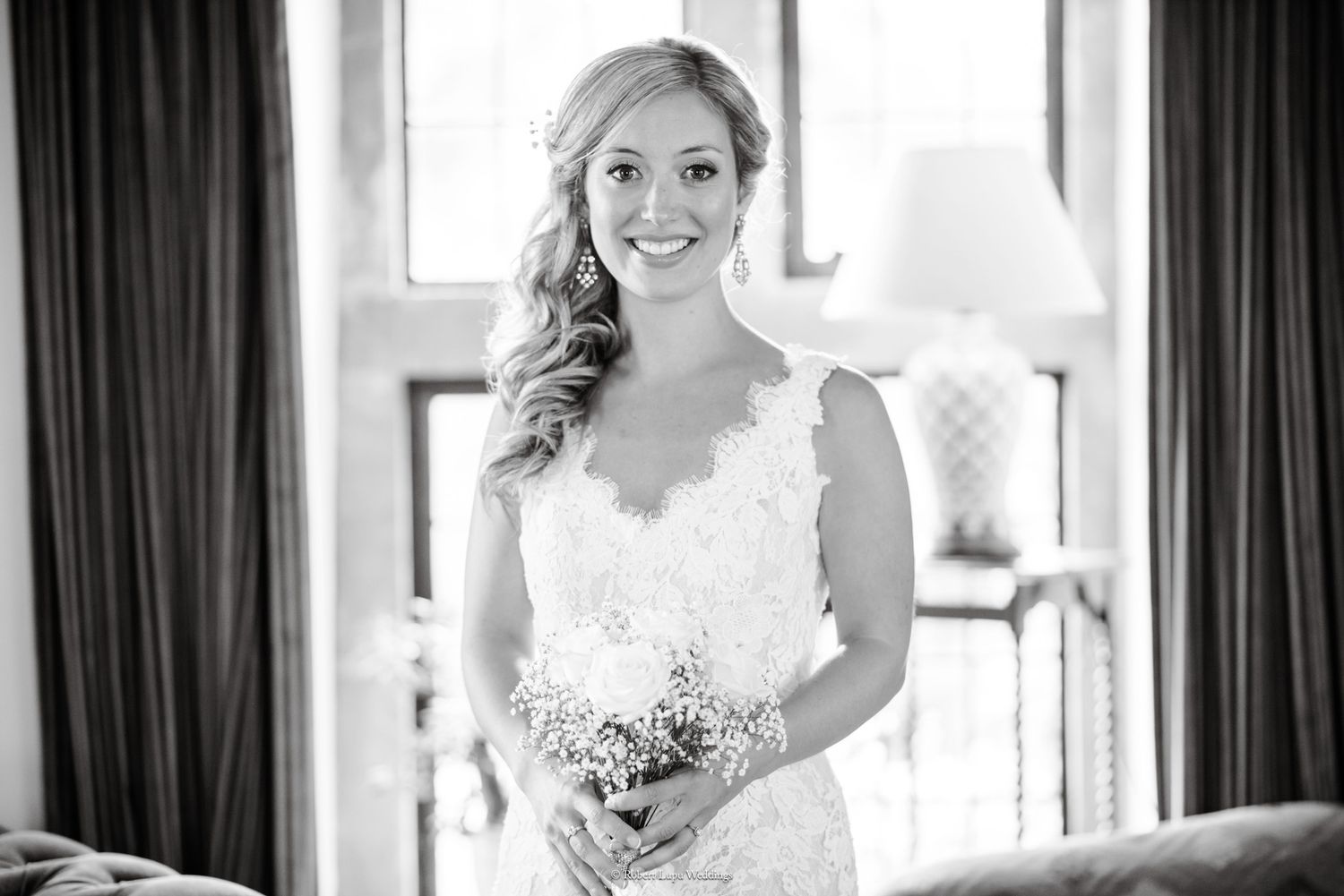 Smiling bride in lace wedding dress holding small bouquet in elegant black and white portrait.