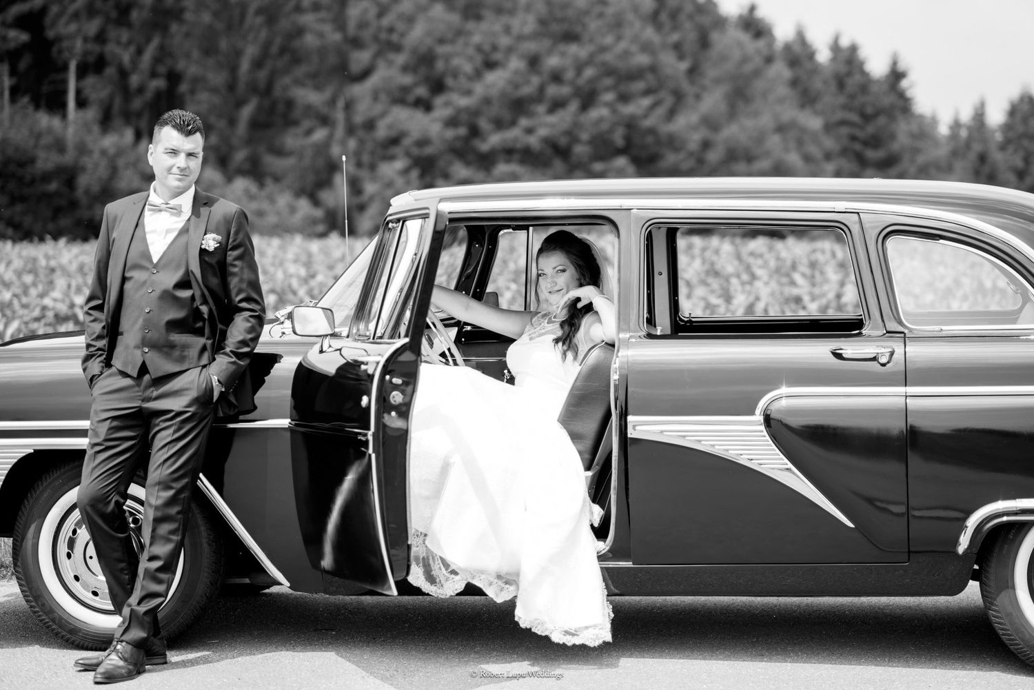 Black and white photo of bride and groom posing with a vintage classic car on their wedding day.