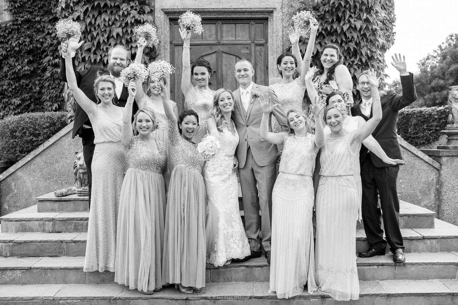 Black and white wedding party photo on steps, bride and groom with bridesmaids and groomsmen celebrating joyfully.
