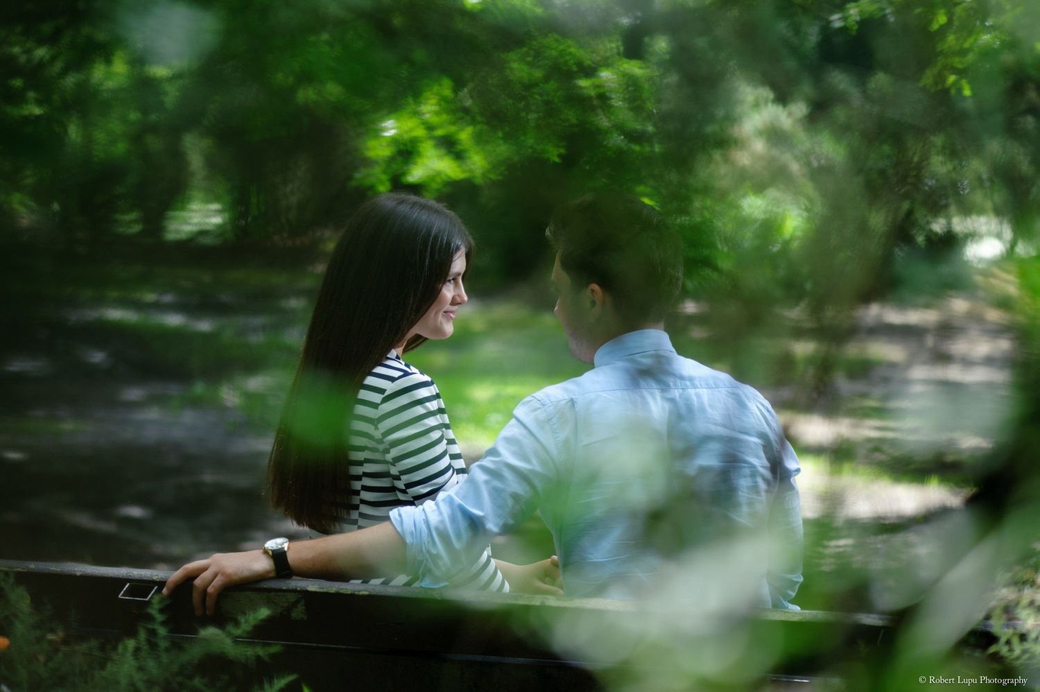 Couple sitting on a bench in a lush green park, smiling and gazing at each other romantically.