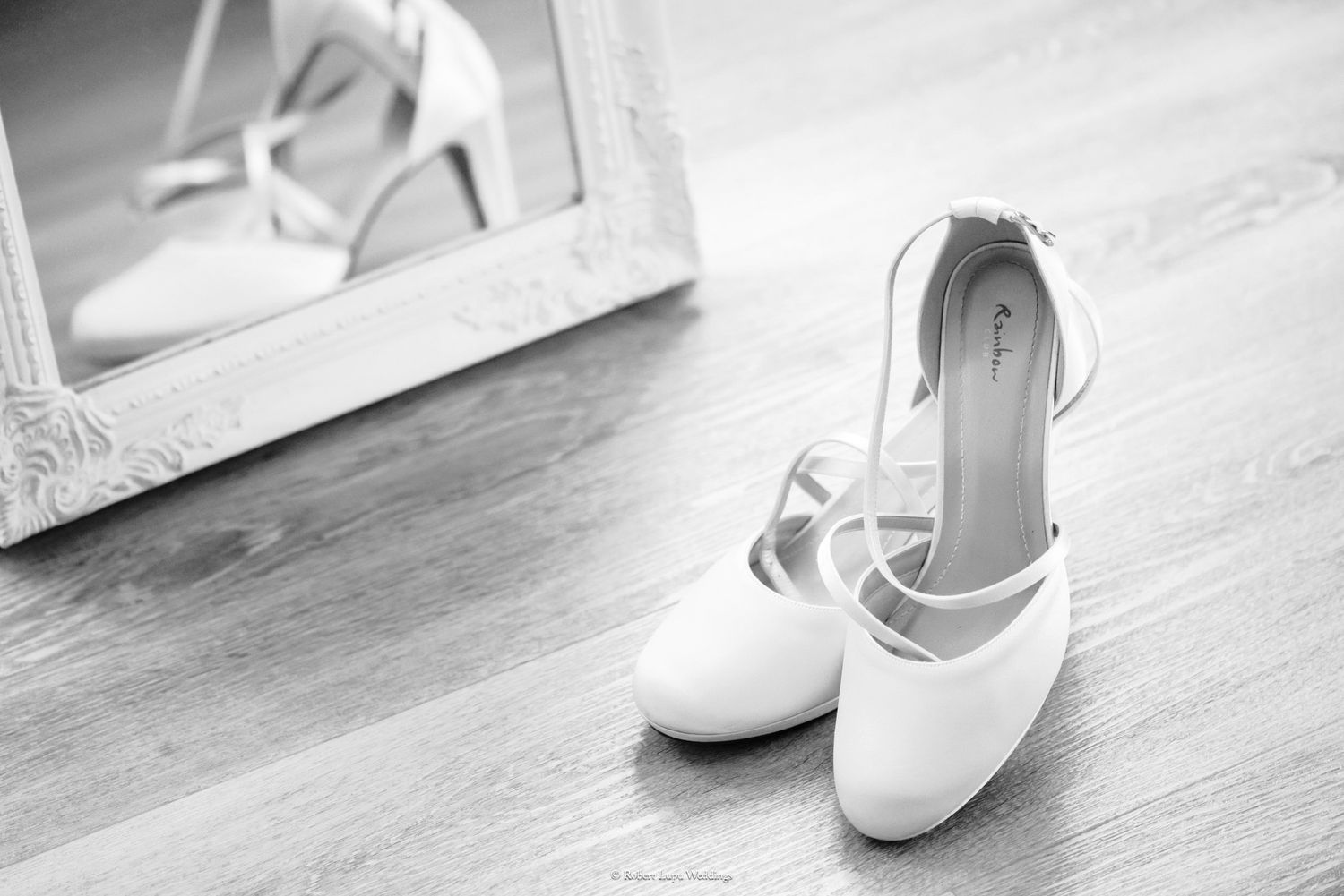 Elegant white bridal heels with ankle straps on hardwood floor beside a mirror in black and white.