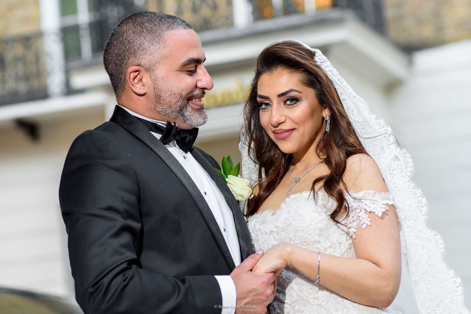 Elegant bride and groom in wedding attire smiling at each other outdoors, bride wearing lace gown and veil.