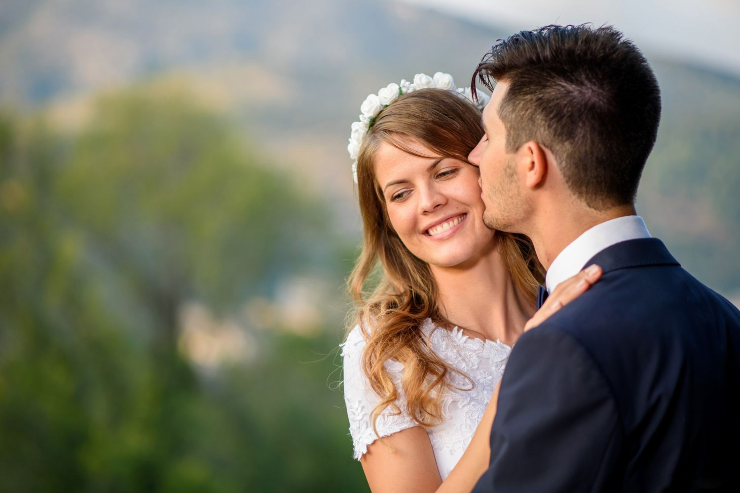 A groom in a navy suit kisses his smiling bride wearing a floral crown on their wedding day outdoors.