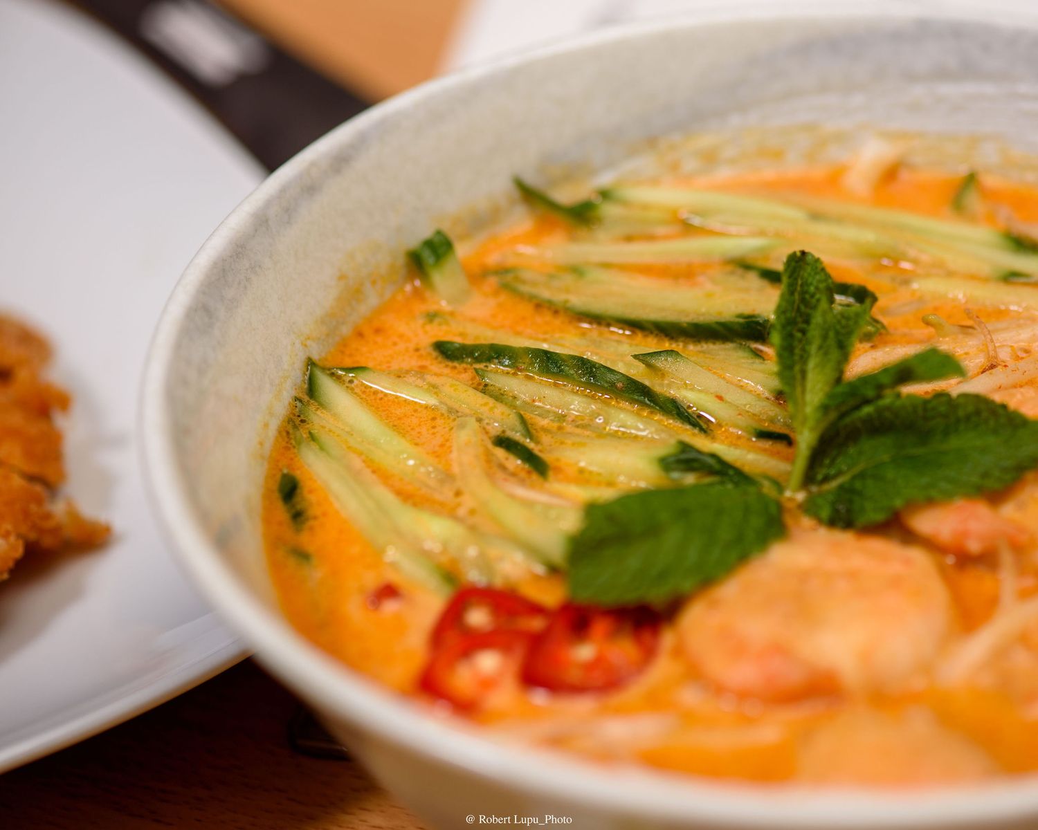 A bowl of creamy red curry soup with shrimp, zucchini strips, fresh mint, and chili peppers served in a ceramic bowl.