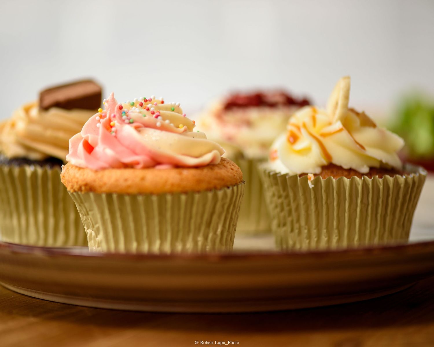 Assorted frosted cupcakes in gold foil liners on a brown plate, topped with colorful sprinkles and decorations.