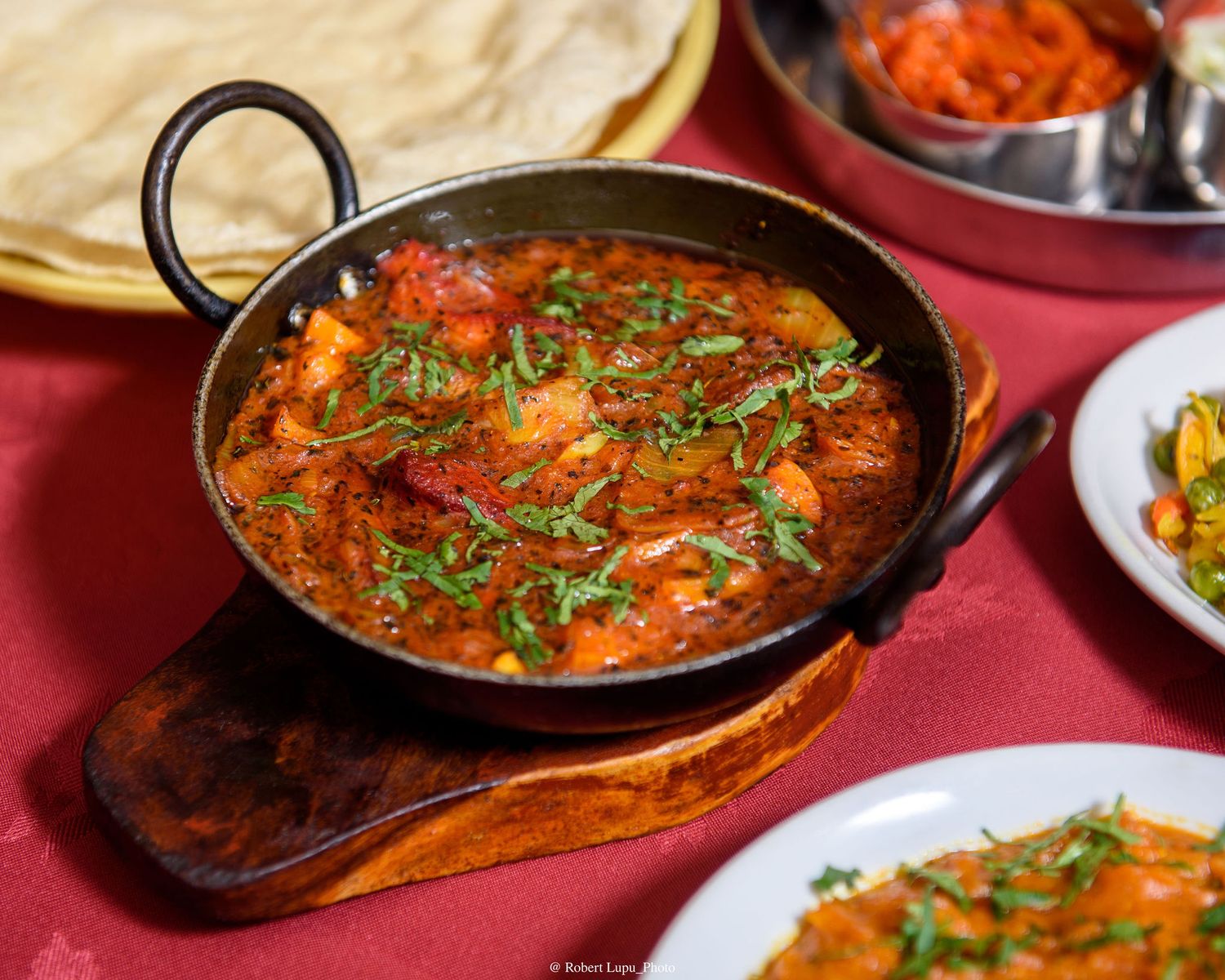 Aromatic Indian curry in a traditional balti dish, garnished with fresh herbs, served with naan bread on a red tablecloth.
