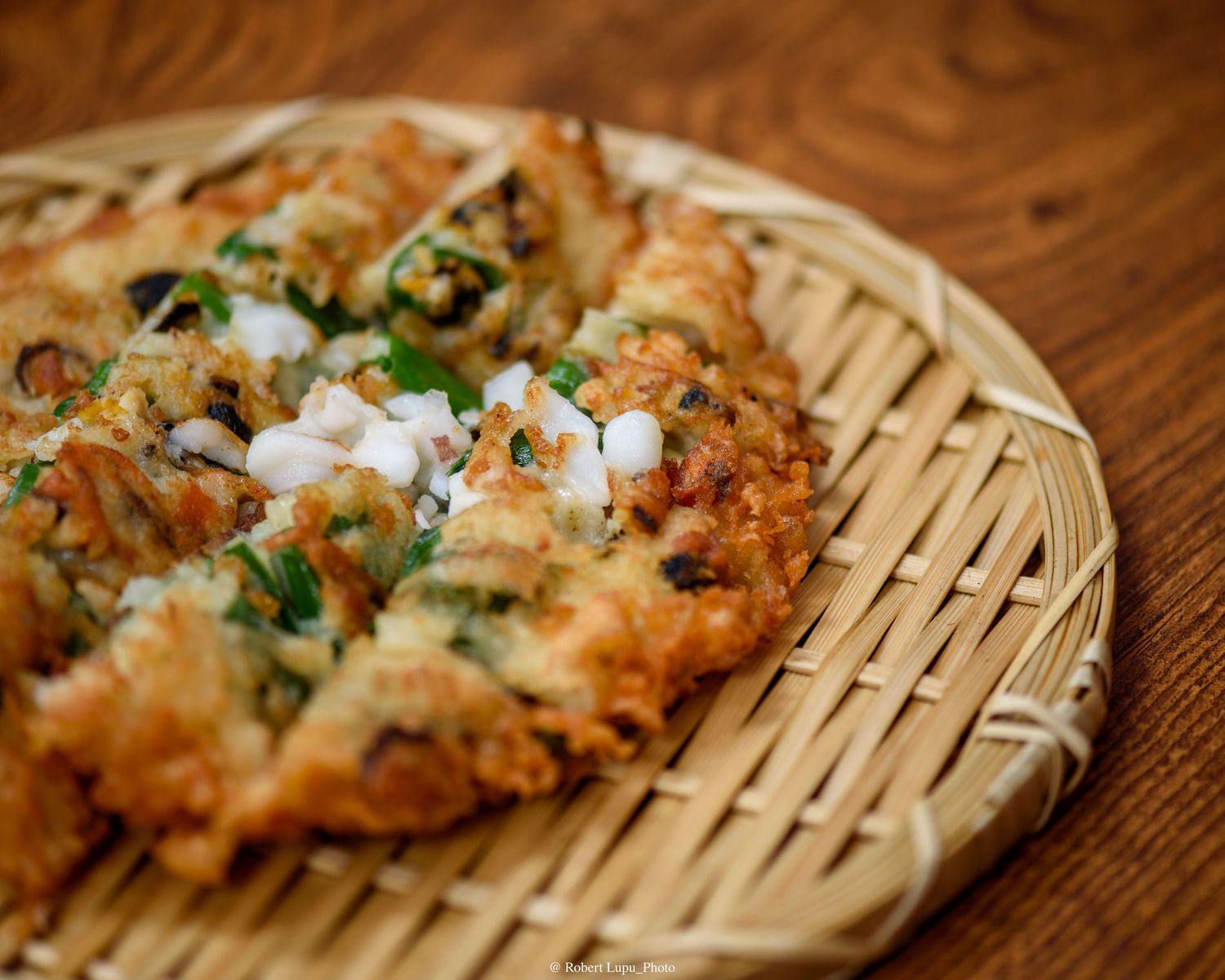 Golden crispy Korean pancake with green onions and toppings served on a woven bamboo tray on wooden table.