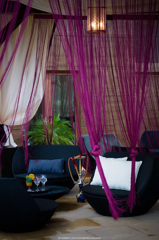 Exotic lounge with vibrant magenta string curtains, dark wicker furniture, white cushions, and a hookah on a black coffee table.