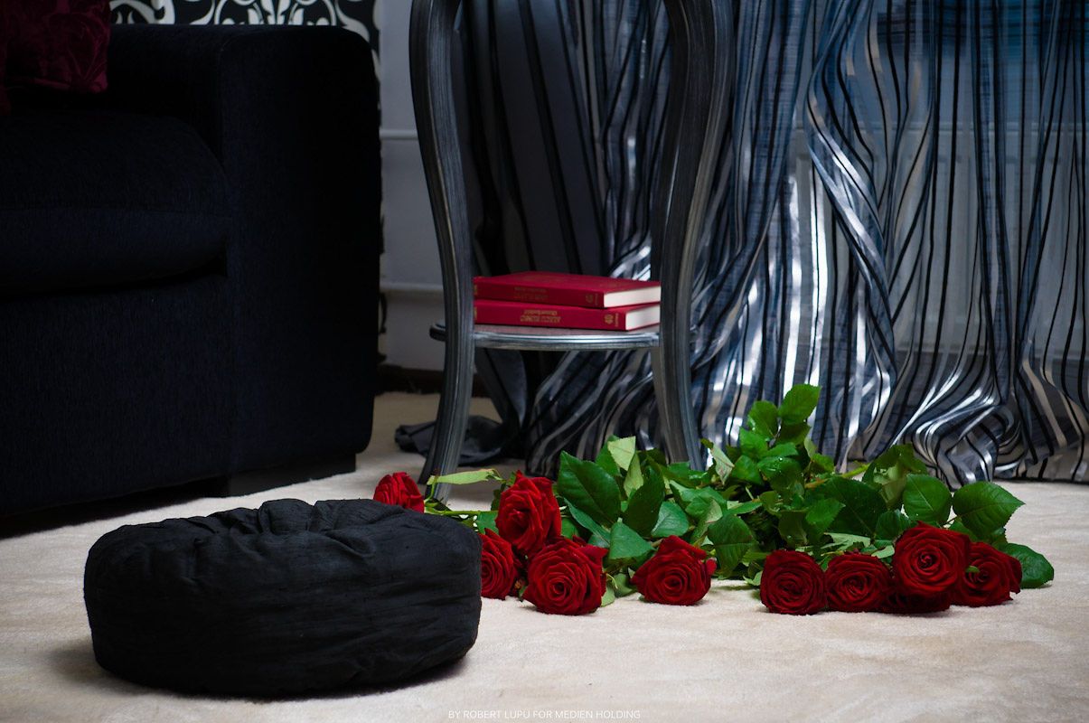 Black quilted hat and red roses bouquet on white carpet with elegant curtains and chair in background.