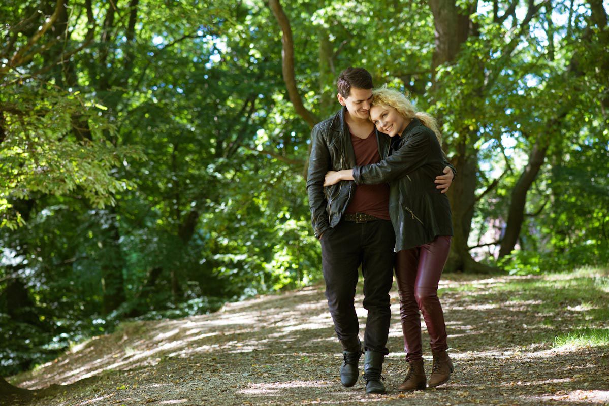Couple walking arm in arm on a tree-lined path, woman leaning on man's shoulder in a lush green forest.