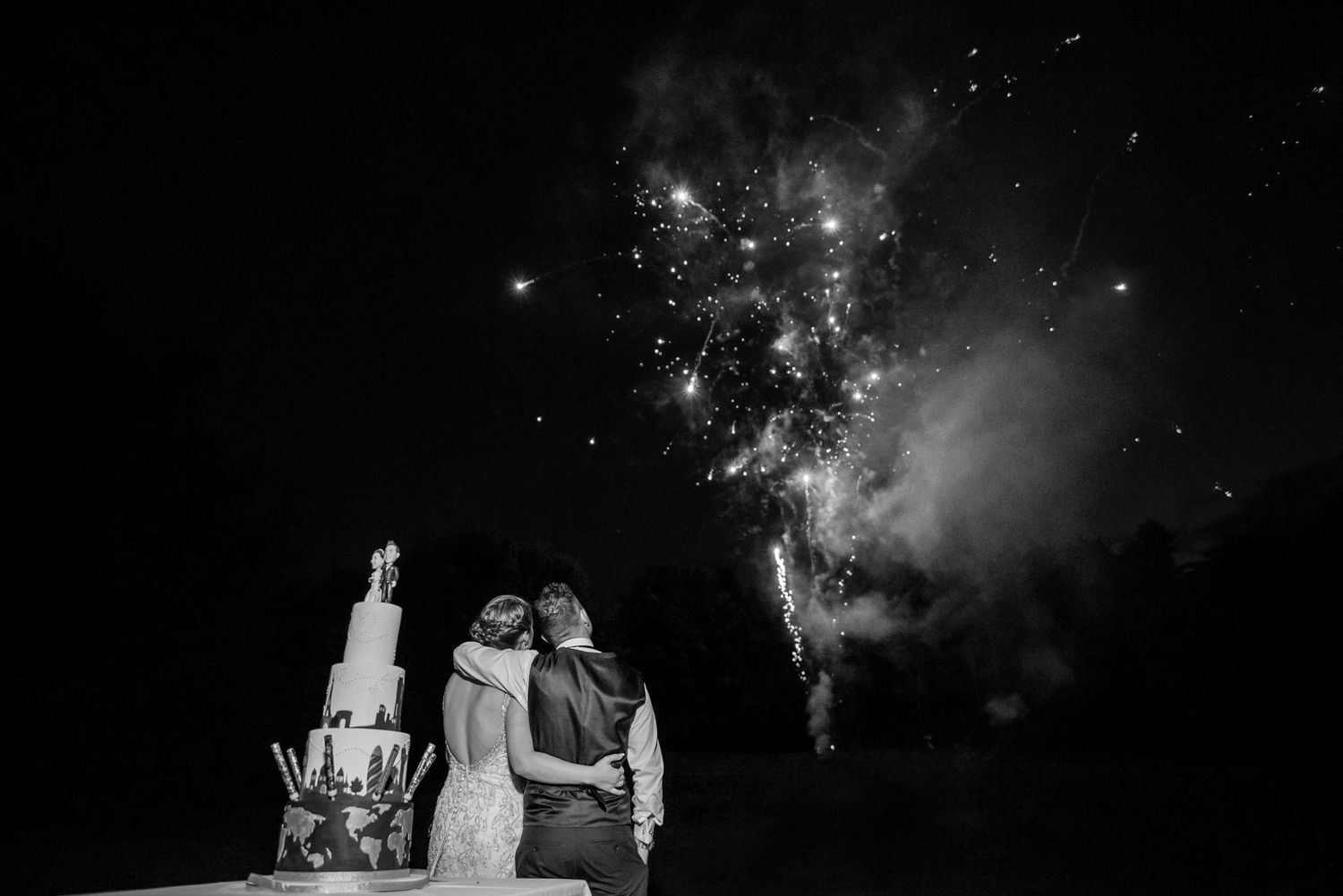 Black and white photo of a couple embracing beside their wedding cake while fireworks illuminate the night sky.