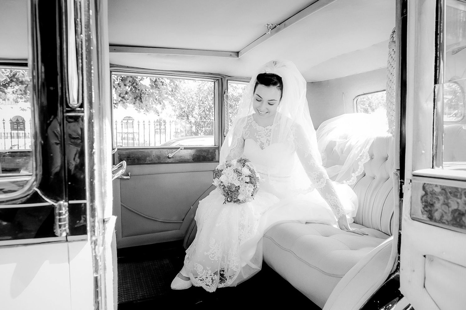 Elegant bride in white gown sitting in a vintage wedding car holding a bouquet.