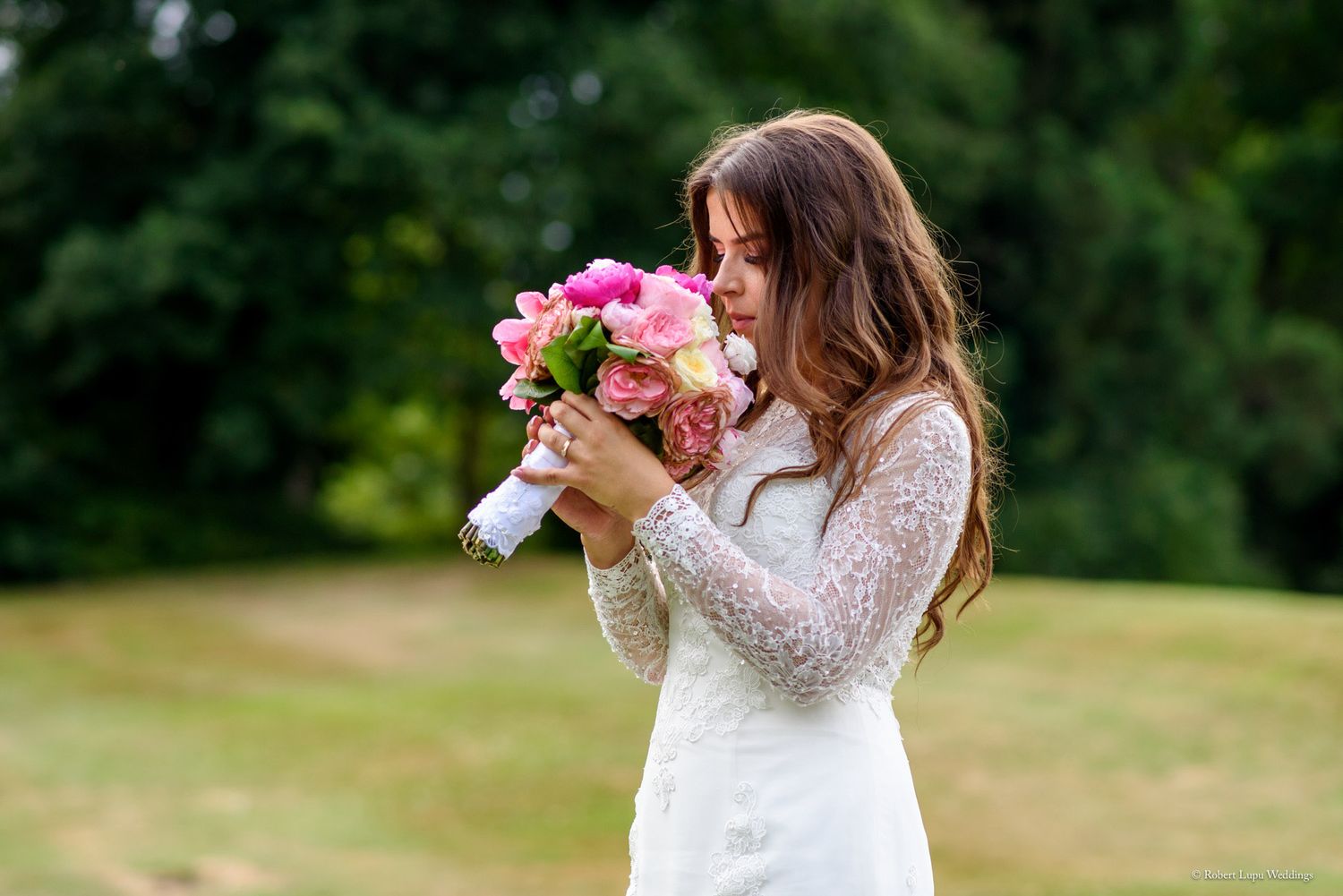 Bride in white lace dress smelling pink peony bouquet outdoors with green trees in background.