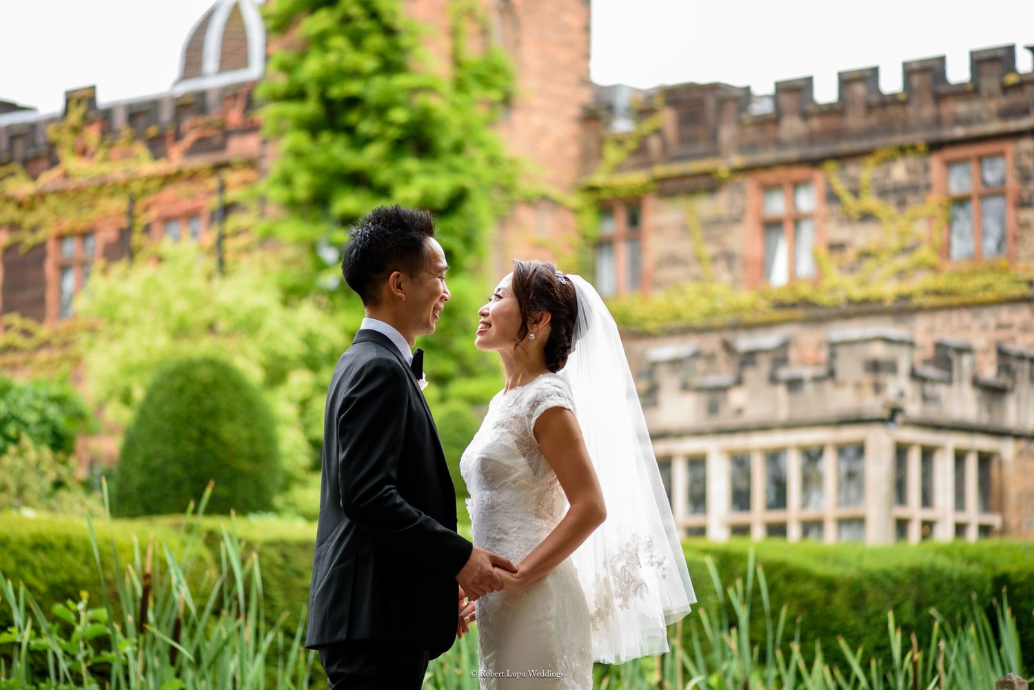 Asian bride and groom holding hands, gazing at each other in lush garden before grand red brick manor house.