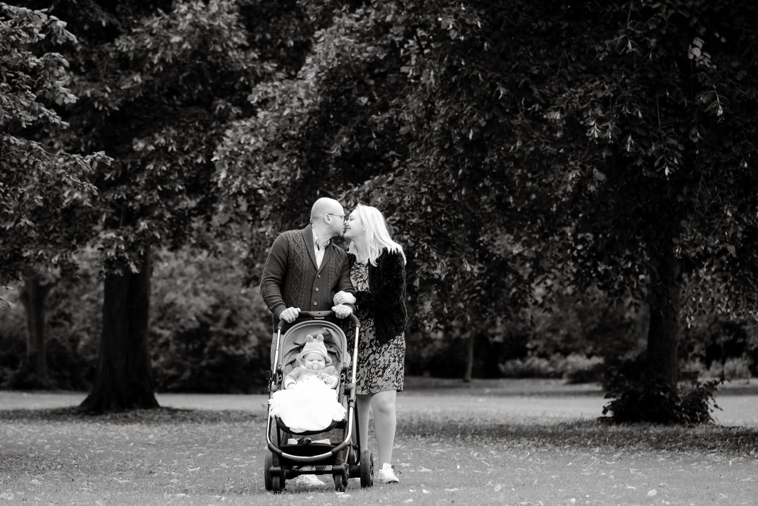 Black and white photo of a couple kissing while pushing a baby stroller through a lush park.