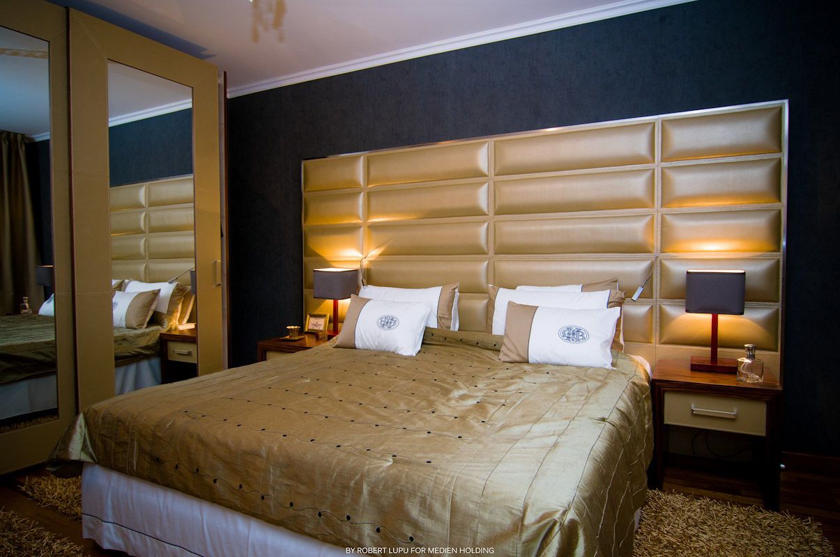 Luxurious hotel bedroom with gold tufted headboard, beige bedding, twin nightstands with lamps, and mirrored wardrobe.