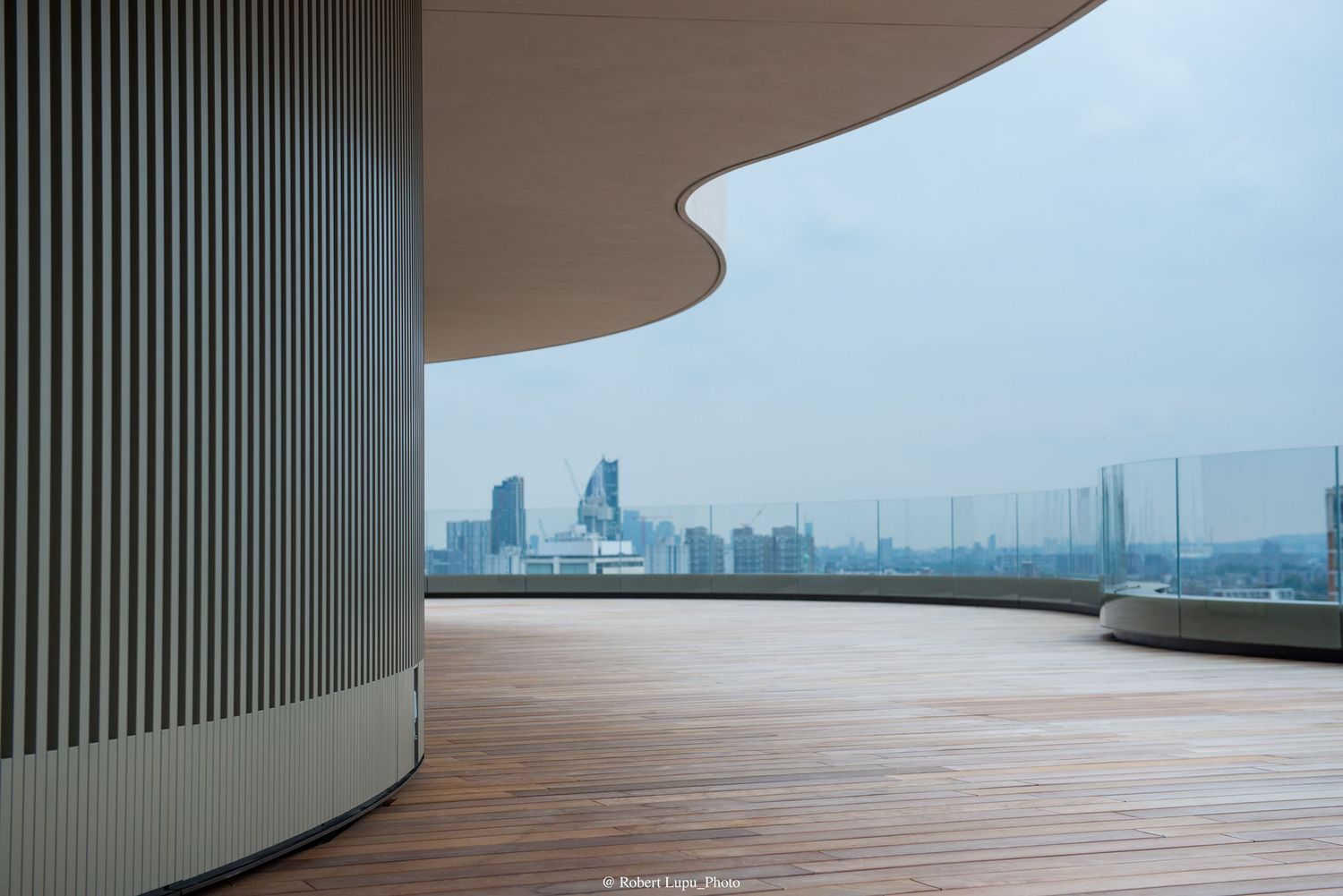 Modern rooftop terrace with wooden deck, curved architecture, and panoramic city skyline views on a hazy day.