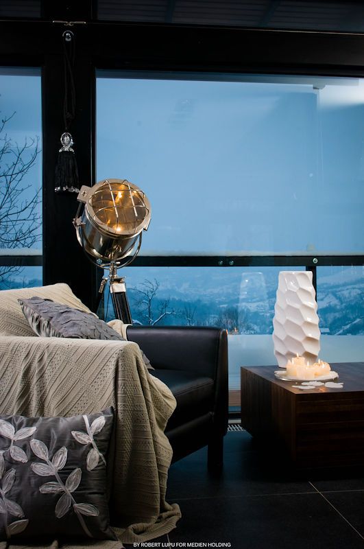 Cozy bedroom with industrial floor lamp, leather sofa, and candles on wooden table against panoramic winter view windows.