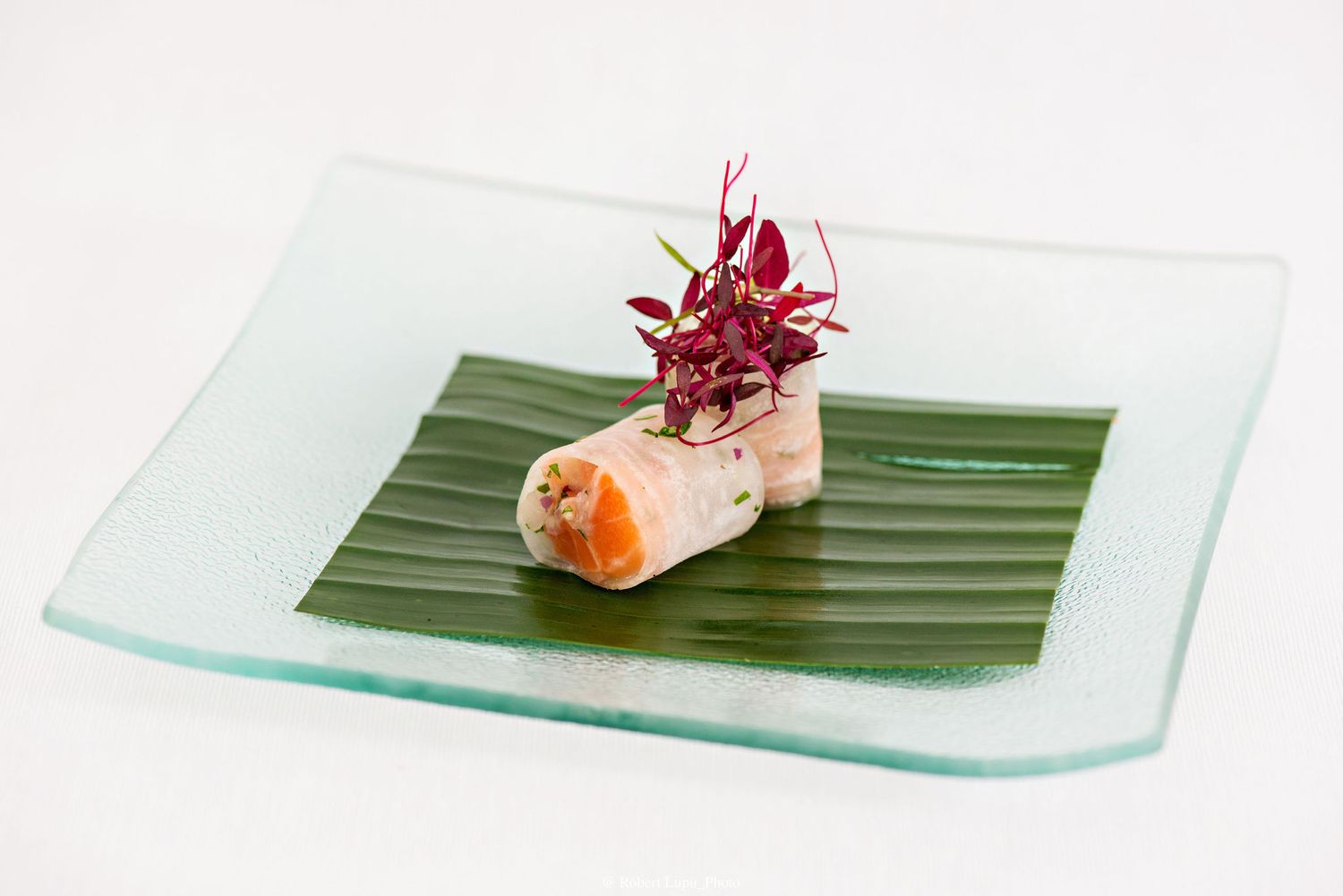 Fresh spring roll with shrimp on banana leaf, garnished with red microgreens, served on a glass plate.