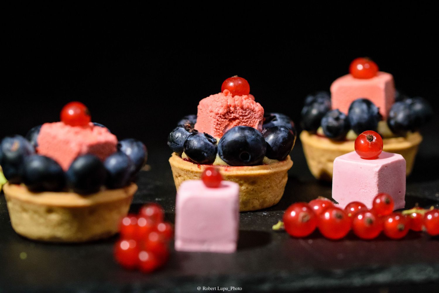 Rustic tartlets topped with blueberries, red currants, and pink ice cream scoops arranged on a dark slate board.