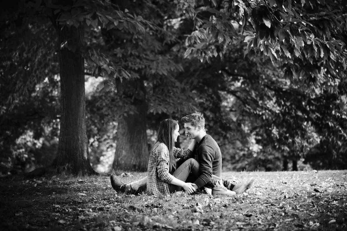 Black and white photo of a couple sitting together on fallen leaves in a park, surrounded by large trees.