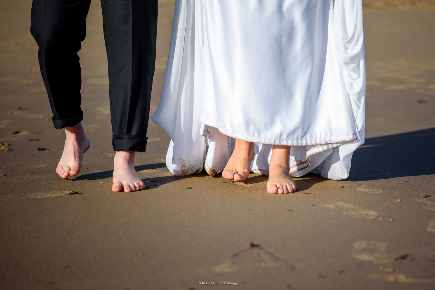 Barefoot bride and groom standing on sandy beach, white wedding dress and black pants visible, romantic outdoor ceremony.