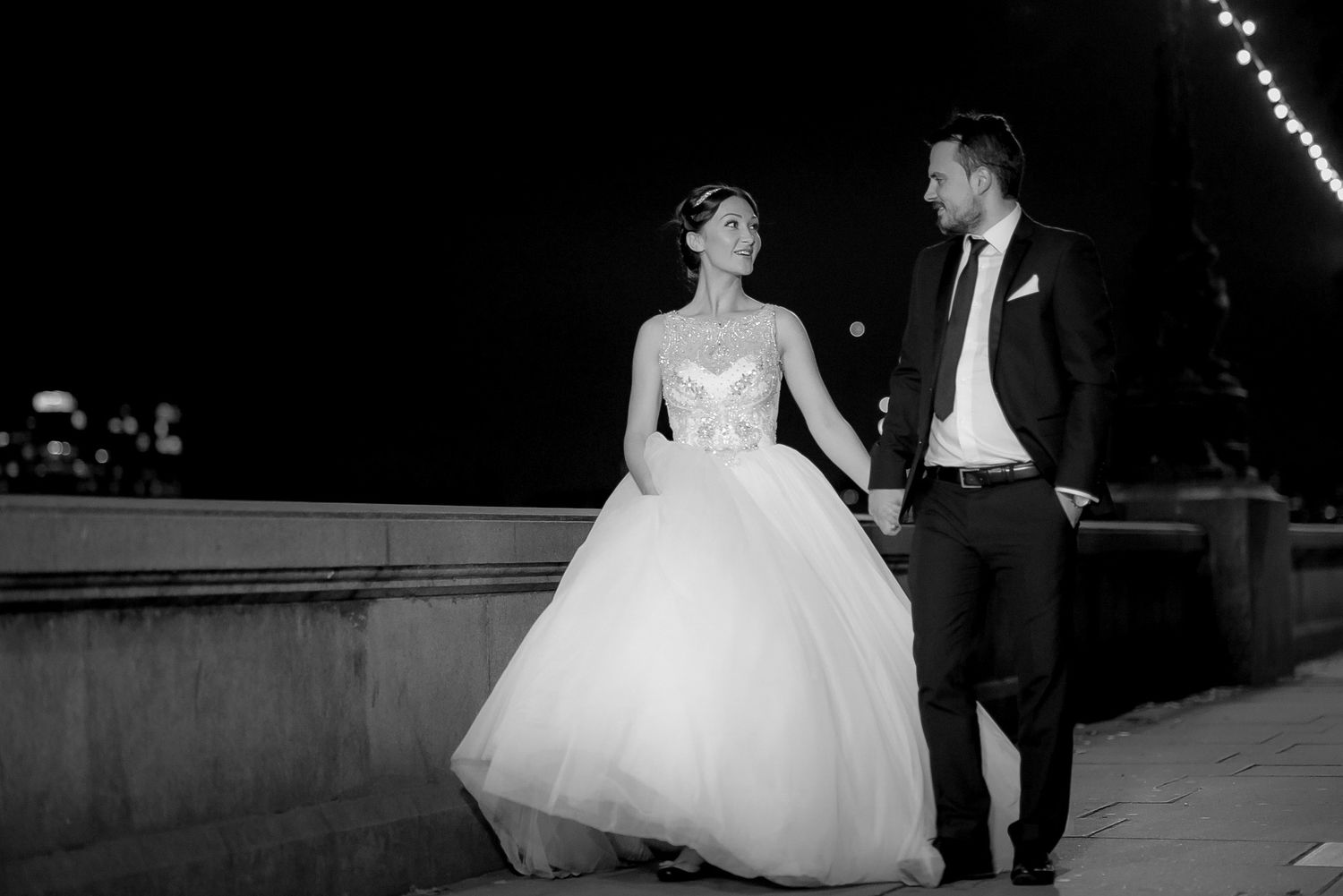 Black and white photo of bride and groom holding hands walking at night with city lights in background.