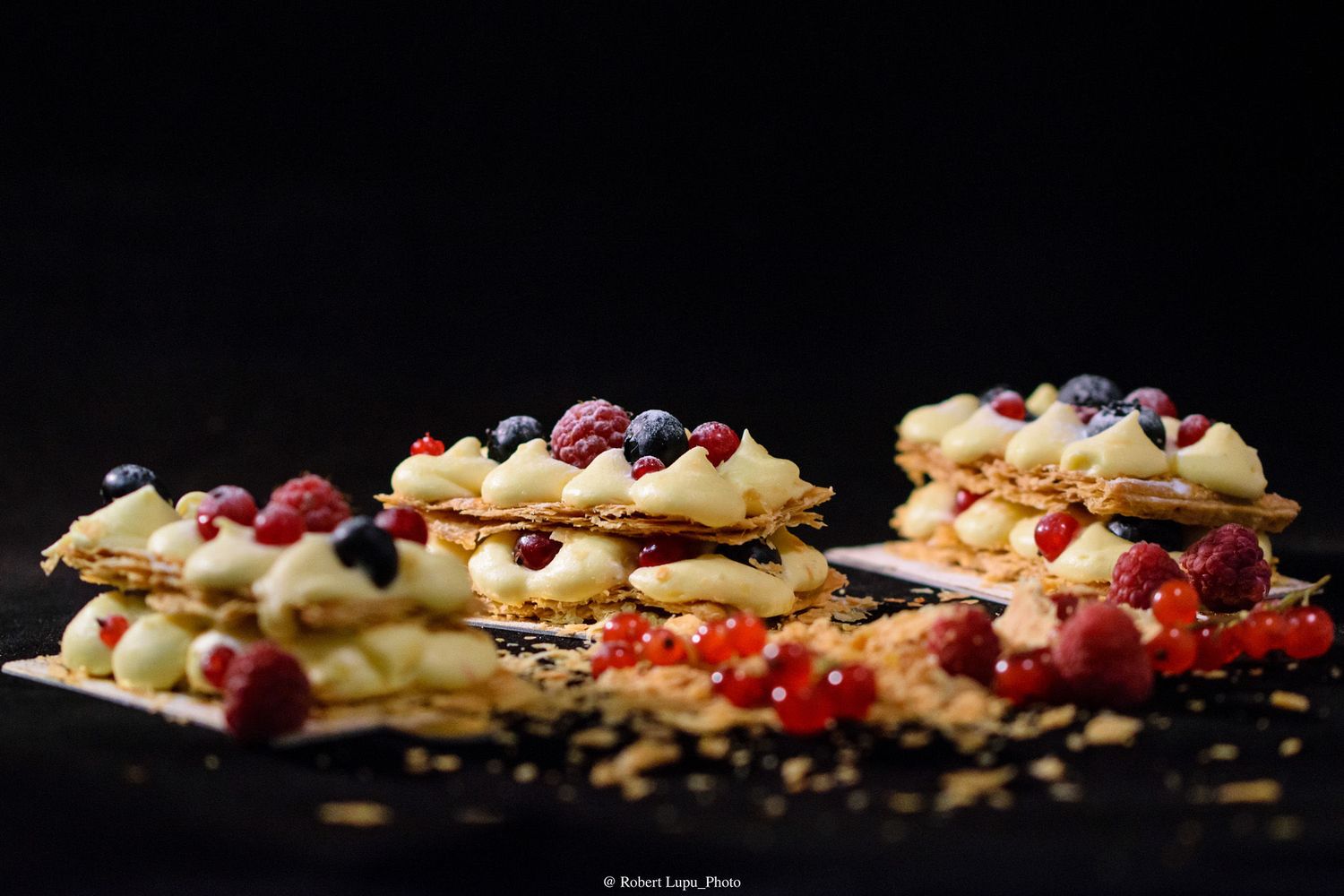 Elegant mille-feuille pastries topped with fresh berries including raspberries and blueberries on a dark background.