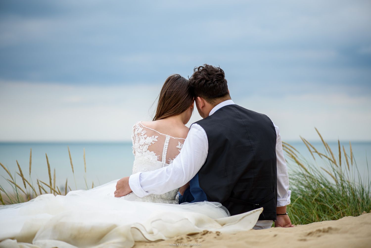 Bride in lace wedding dress and groom in black vest sit together on sandy beach dunes overlooking calm ocean.
