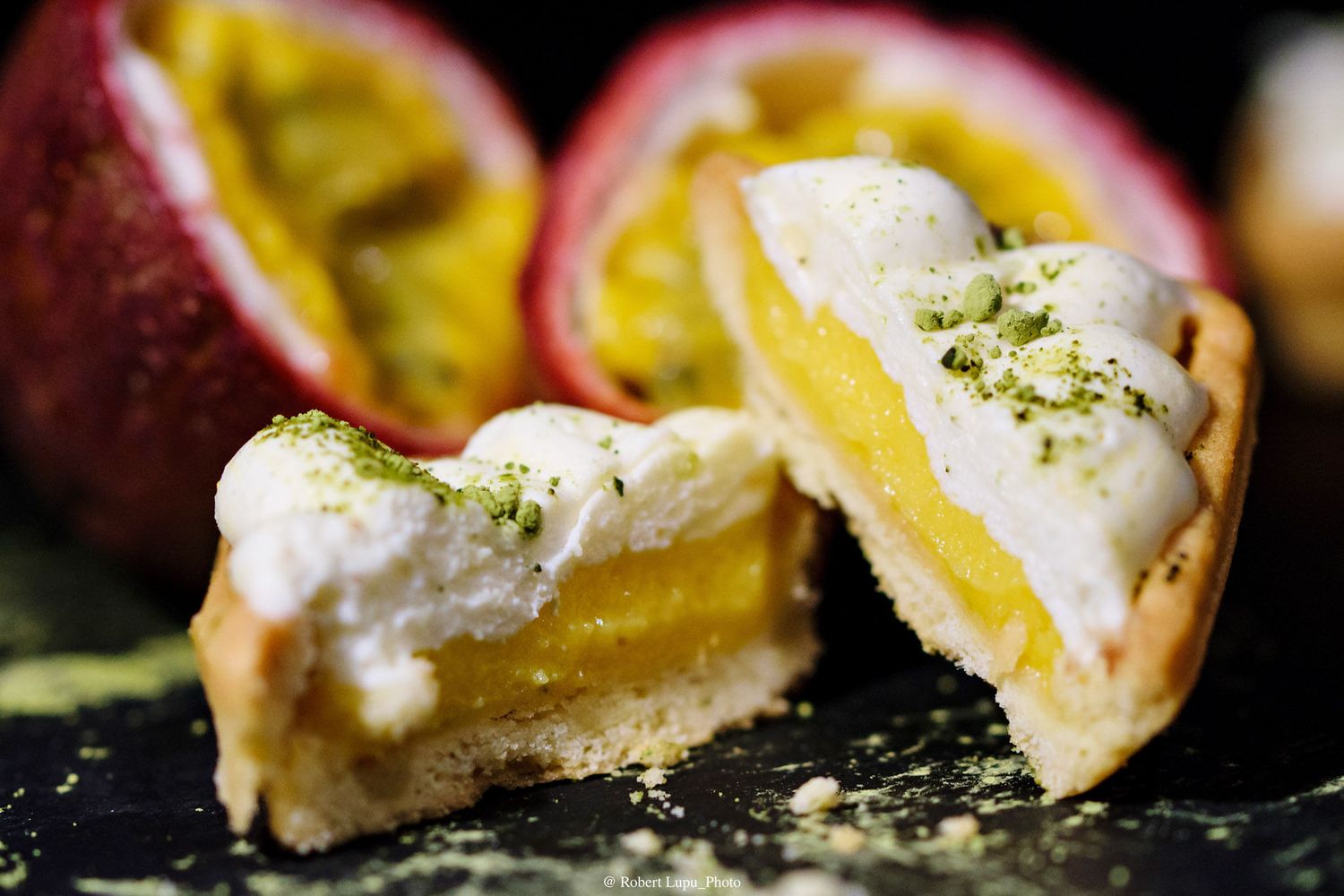 Passion fruit tart cut in half revealing creamy yellow filling topped with white meringue and green zest on dark surface.