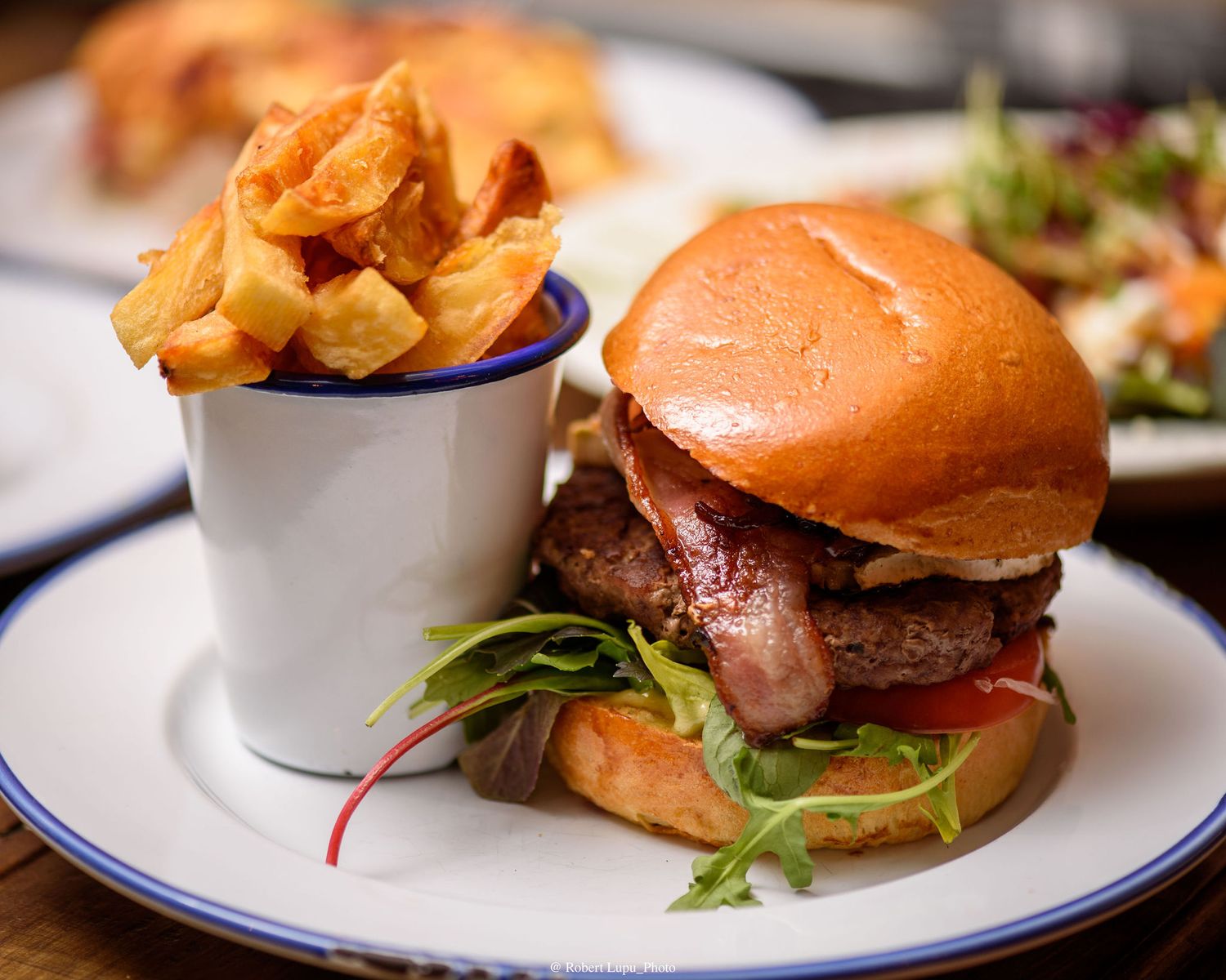 Juicy beef burger with bacon and greens on a brioche bun, served with crispy thick-cut fries in a white enamel cup.