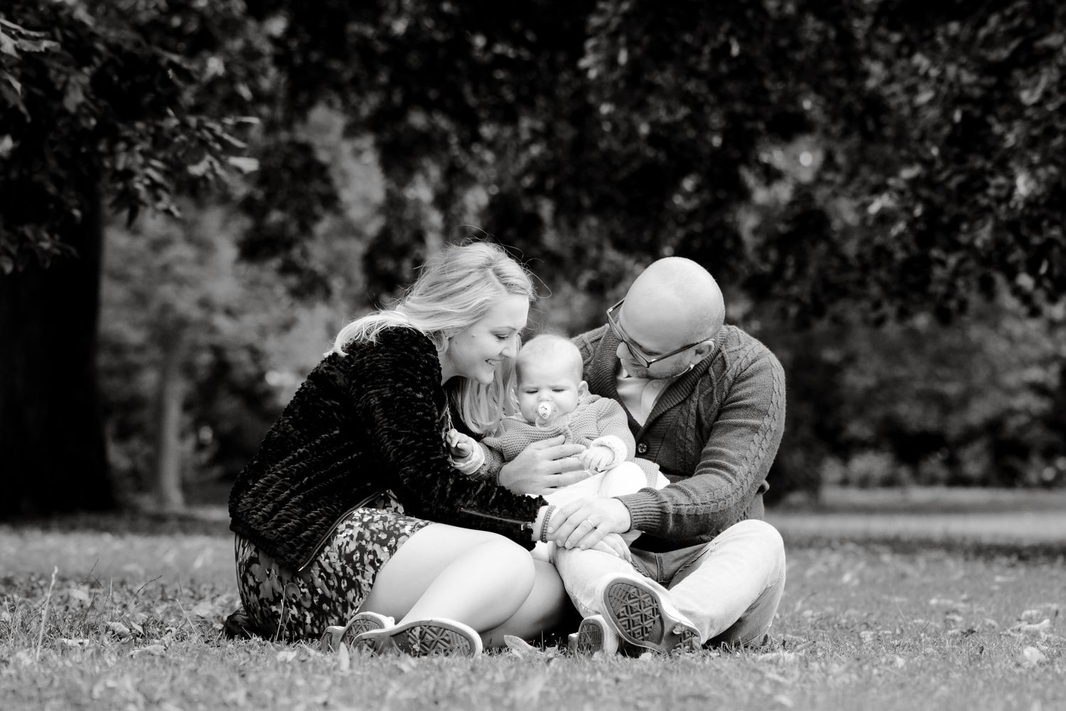 Black and white photo of parents lovingly kissing their baby while sitting together on grass in a park.