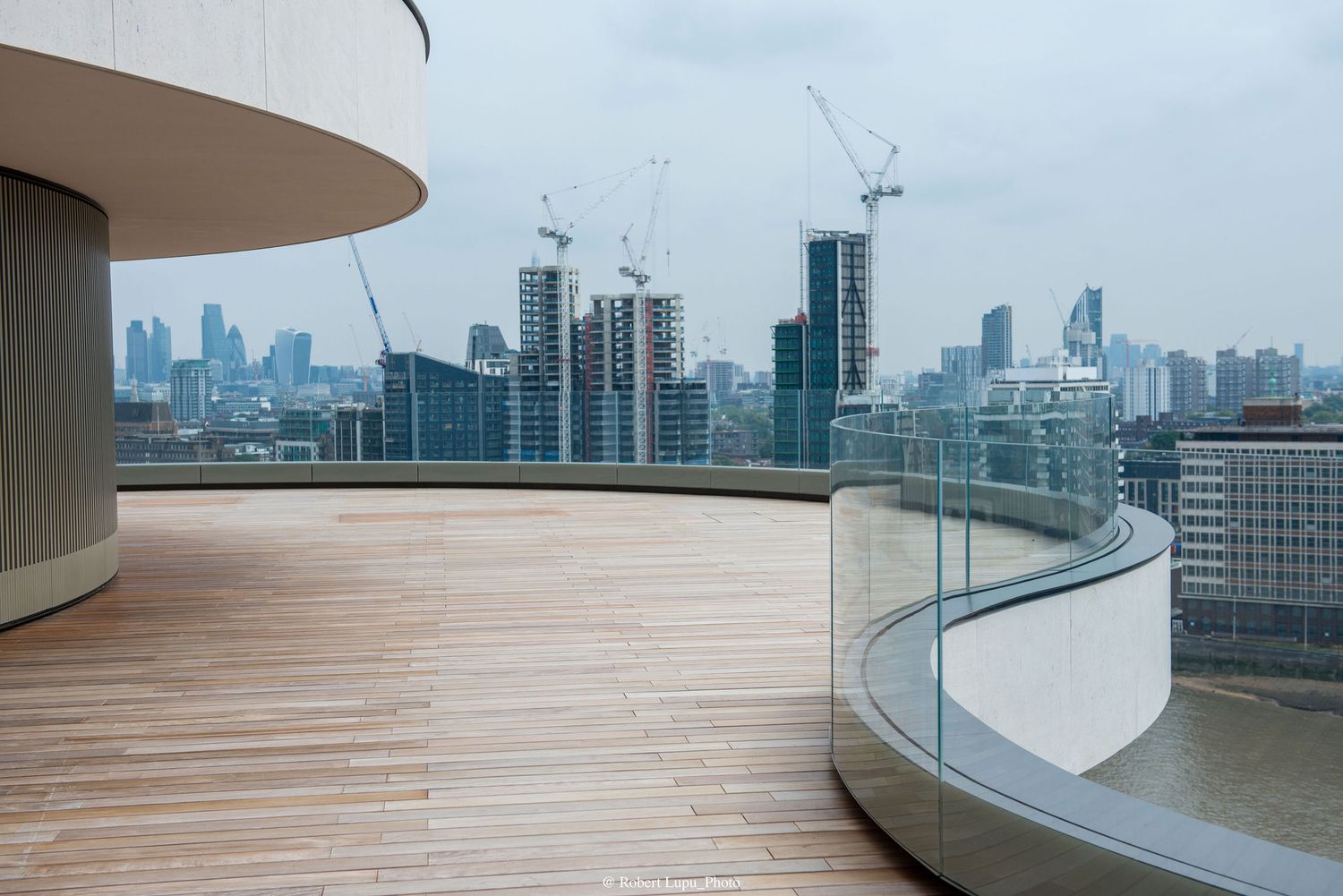 Modern rooftop terrace with wooden deck and glass railing overlooking London skyline with cranes and city buildings.