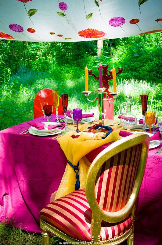 Vibrant outdoor dining setup with fuchsia tablecloth, gold striped chair, colorful candles, and floral decorations.