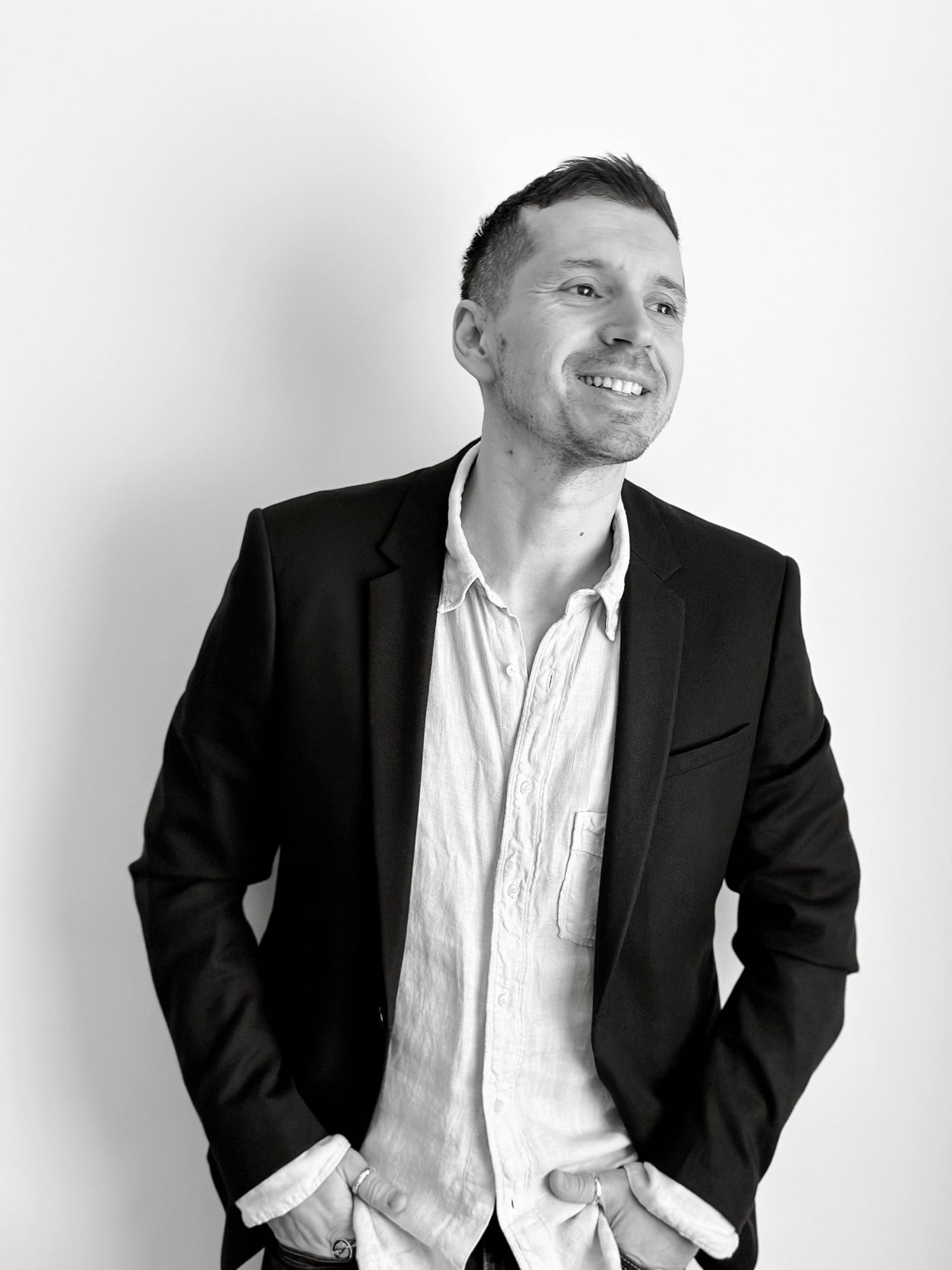 Black and white portrait of a smiling man in a blazer and open-collar shirt, hands in pockets.