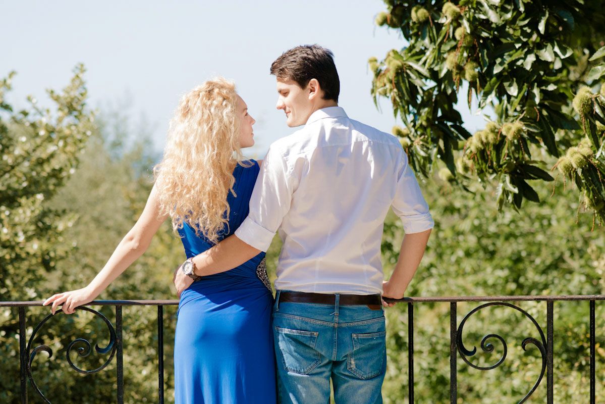 Romantic couple standing on a balcony surrounded by lush green trees, woman in blue dress embracing man in white shirt.