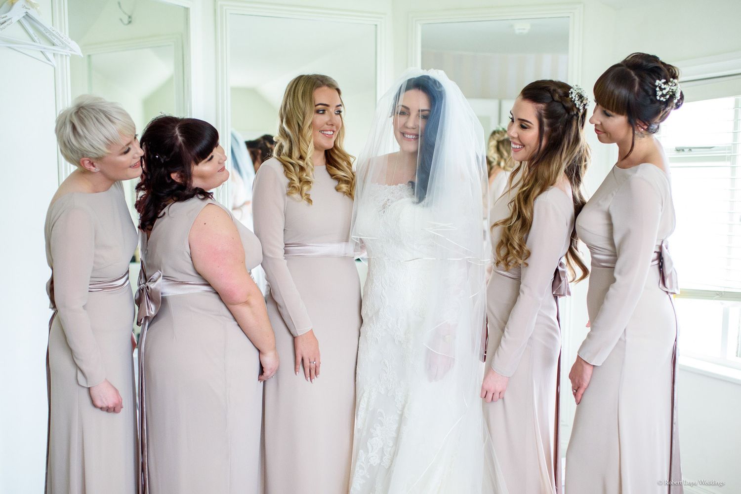 Bride in white gown and veil stands with five bridesmaids in taupe dresses, sharing a joyful moment together.