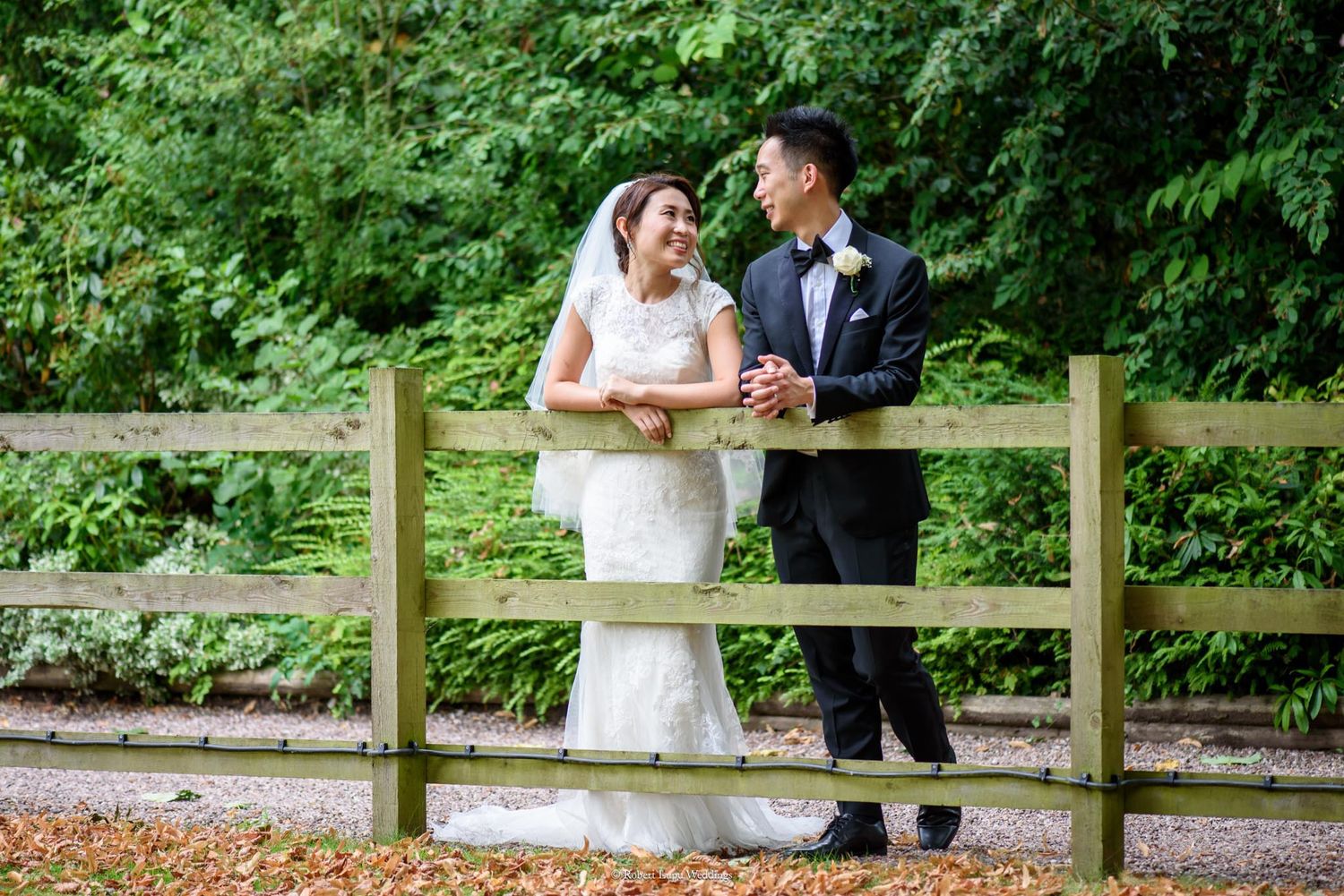 A bride and groom smile at each other leaning on a wooden fence surrounded by lush green trees.