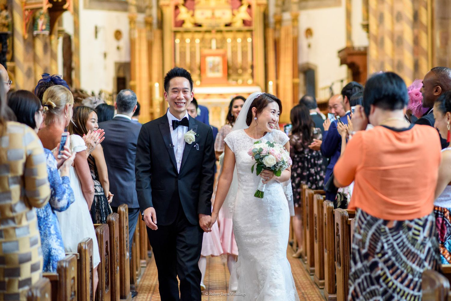 Newlywed Asian couple walking down church aisle after wedding ceremony, bride in white gown holding bouquet.