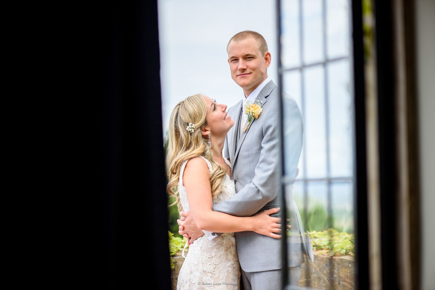 Bride in lace gown embraces groom in gray suit with yellow boutonniere, sharing a romantic moment at their outdoor wedding.