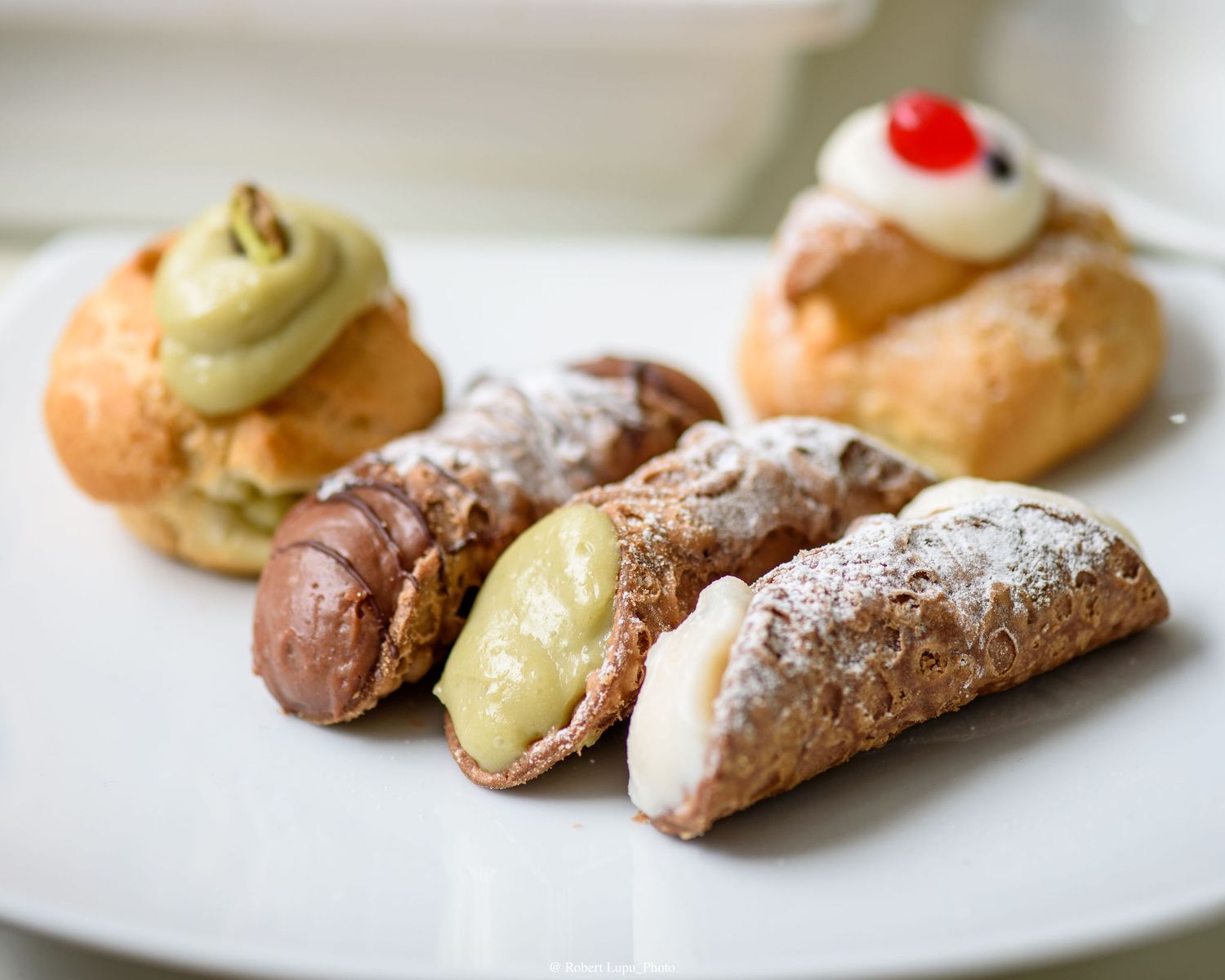 Assorted Italian pastries including cannoli dusted with powdered sugar and cream puffs topped with whipped cream on a white plate.